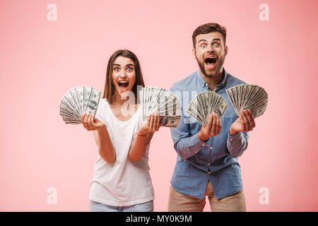 Portrait of a joyful young couple holding bunch of money banknotes and celebrating success isolated over pink background Stock Photo