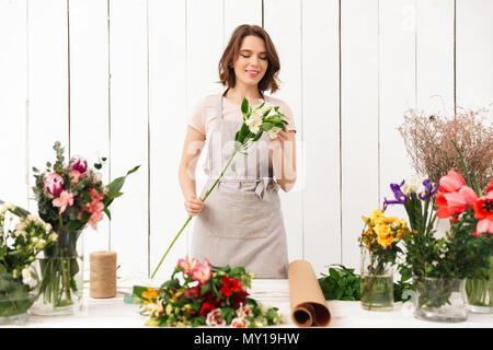 Young pretty smiling florist woman standing near table with different flowers and looking aside in workshop. Stock Photo