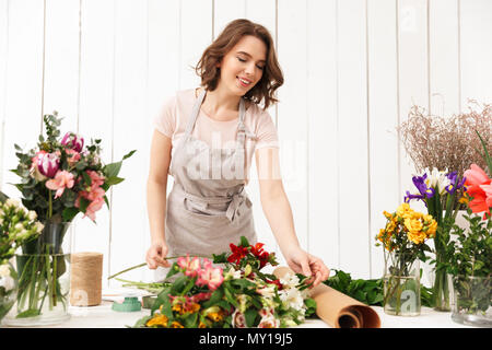 Young pretty smiling florist woman standing near table with different flowers and looking aside in workshop. Stock Photo