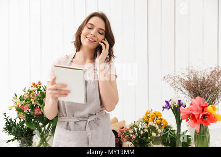 Young florist woman standing near table with different flowers in studio and calling to clients with notes in hand Stock Photo