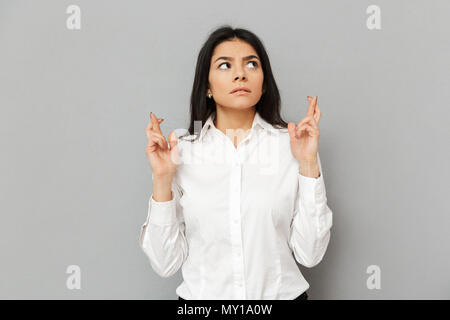 Portrait of a worried young businesswoman holding fingers crossed for good luck isolated over gray background Stock Photo