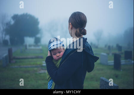 A grieving young mother is standing with her baby by a tombstone in a graveyard Stock Photo