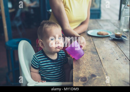 A mother is helping her baby drink from a cup by the window in a cafe Stock Photo