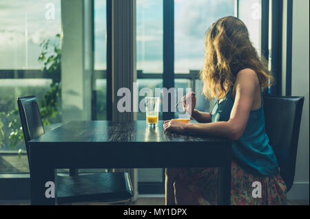 A young woman is sitting at a table by the window in a modern city apartment and is having breakfast Stock Photo