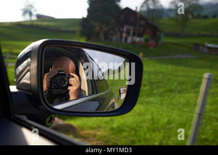 Close up of a photographer with a DSLR camera taking a photo of himself in the outside car mirror whilst sitting in the car on the way to the mountain Stock Photo