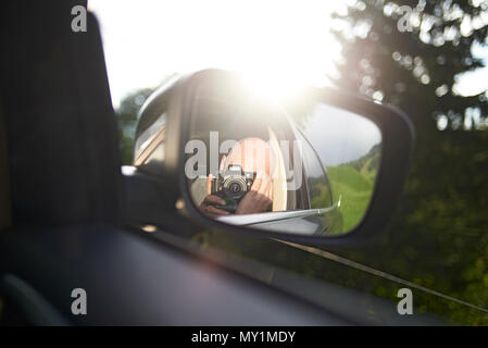 Close up of a photographer with a DSLR camera taking a photo of himself in the outside car mirror whilst sitting in the car on the way to the mountain Stock Photo