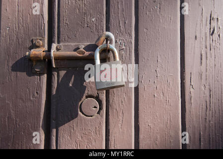 A rusty slide bolt secured with a Lockwood padlock and a embedded key lock in an old wooden door with peeling paint Stock Photo