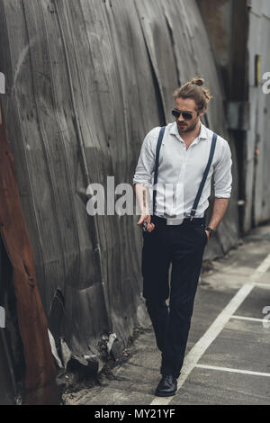 Handsome bearded stylish man in formal wear and sunglasses holding lighter and walking with hand in pocket Stock Photo