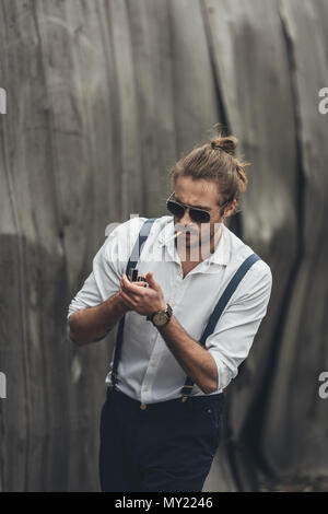 Portrait of handsome stylish bearded man in sunglasses and suspenders lighting cigarette with lighter Stock Photo