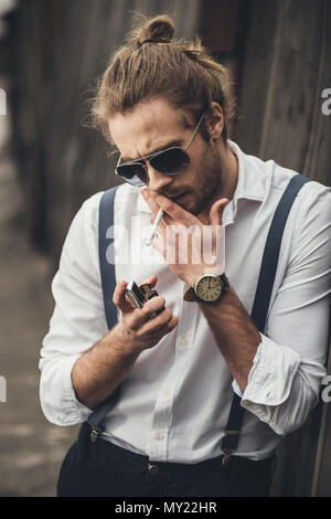 Portrait of bearded handsome man in sunglasses lighting cigarette with lighter Stock Photo