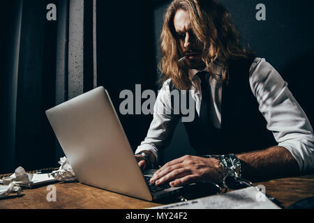 Stylish long haired man smoking cigarette and using laptop at table Stock Photo