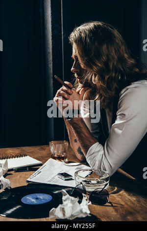Handsome young bearded man smoking cigar while sitting at table with newspaper, ashtray, vinyl record and glass of whisky Stock Photo