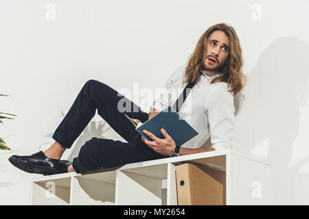 Handsome young long haired businessman lying on bookshelf and reading book in office Stock Photo