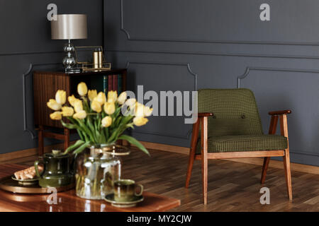 Real photo of a green, old-fashioned armchair in living room interior with a wooden cupboard in the background and blurred yellow tulips in the foregr Stock Photo