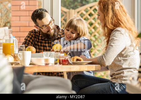 Happy boy holding a citrus, sitting between mother and father. Lovely family eating breakfast Stock Photo