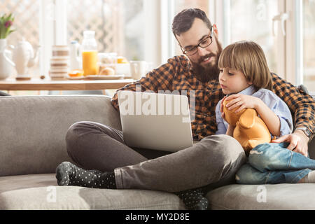 Kid with toy sitting next to his father and watching a movie on a laptop Stock Photo