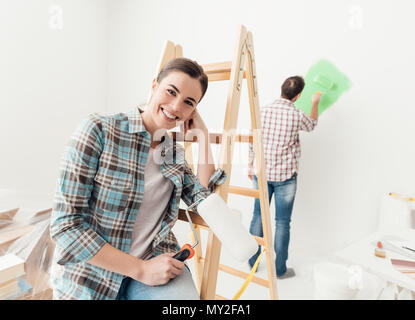 Home makeover and renovation: young couple painting their new house, the man is using a paint roller and the woman is leaning on a ladder and smiling Stock Photo