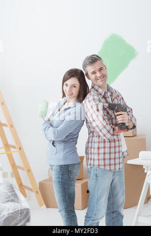 Smiling loving couple doing home renovations, the woman is holding a paint roller and the man is using a drill Stock Photo