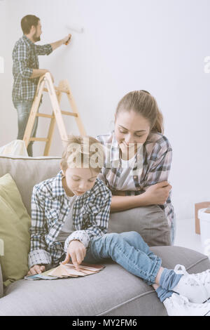 Happy young family renovating and decorating their home, the boy is picking colors swatches with his mother and his father is painting on the backgrou Stock Photo