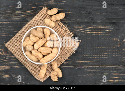 Full ceramic bowl of peanuts in the shell on sackcloth and empty wooden background, top view Stock Photo