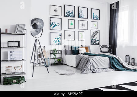 Real photo of a cozy bed standing next to a black lamp in a monochromatic bedroom with shelves with ornaments and posters on a wall Stock Photo