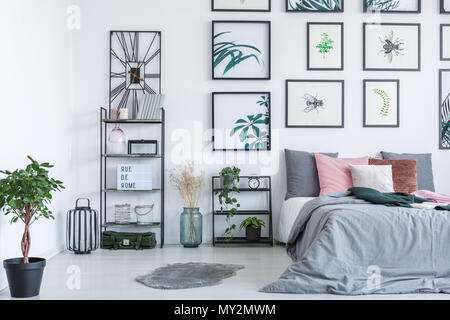 Real photo of a bed standing next to the shelves with ornaments and plants in bedroom interior with paintings on a wall Stock Photo