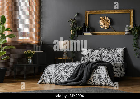 Glass lamp placed on a gold end table standing by the king-size bed in dark grey bedroom interior with windows and green plants Stock Photo