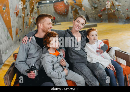 A family with kids at gym, sitting and relaxing on a bench, with climbing walls in the background