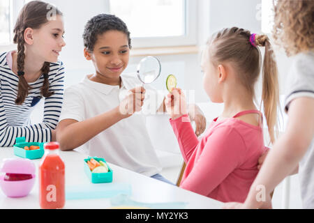 African-american boy with magnifying glass looking at cucumber slice at school Stock Photo