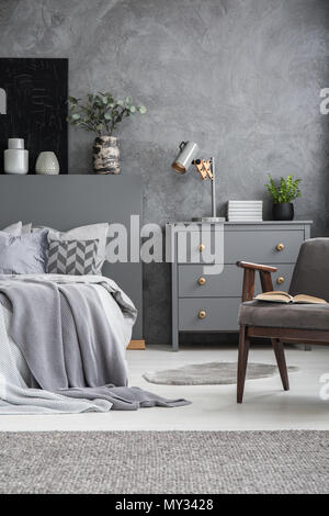 Armchair next to bed in grey bedroom interior with cabinet against concrete wall. Real photo Stock Photo