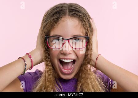 Teen girl with glasses, with long hair scratches her head and is emotional puzzled. Pink studio background Stock Photo