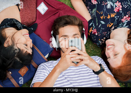 Young man lying on grass with friends, using smartphone and listening to headphones Stock Photo
