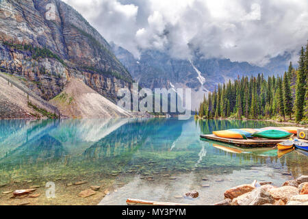 Reflective Moraine Lake at Lake Louise near Banff in the Canadian Rockies, with canoes on nearby dock and heavy clouds descending on the Valley of Ten Stock Photo