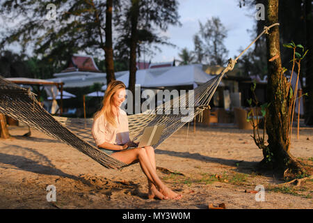 Young woman using laptop and sitting on sand in wicker hammock. Stock Photo