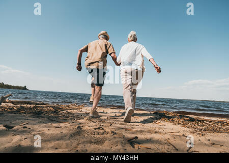Back view of senior couple holding hands and running on sandy beach Stock Photo