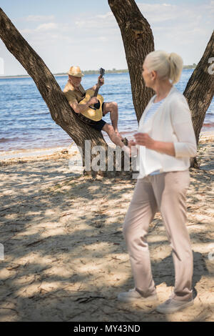 Beautiful senior woman standing on sandy beach while senior man playing guitar behind Stock Photo