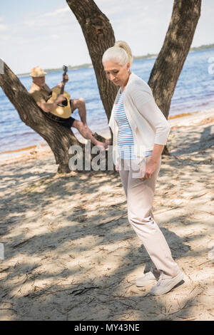 Beautiful senior woman standing on sandy beach while senior man playing guitar behind Stock Photo