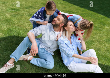 Happy family sitting on grass with american flag, celebrating 4th july - Independence Day Stock Photo