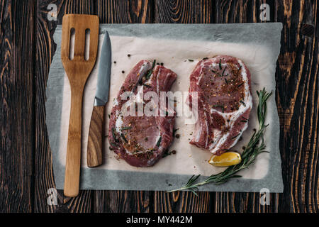 top view of two raw ribeye steaks with spices and rosemary on baking paper on wooden tabletop Stock Photo