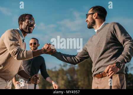 happy stylish multicultural friends shaking hands while playing golf on golf course Stock Photo