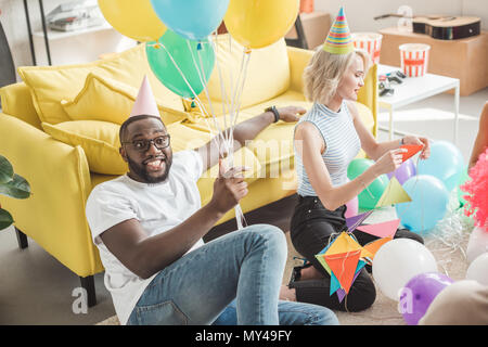stylish african american man in eyeglasses and party hat holding colorful balloons in hand and young woman with party garlands sitting near Stock Photo