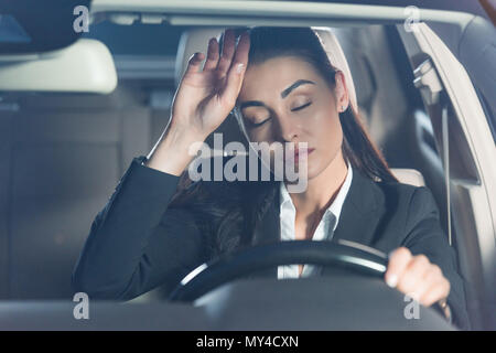 Young tired woman in suit sitting in driver seat and touching her forehead Stock Photo
