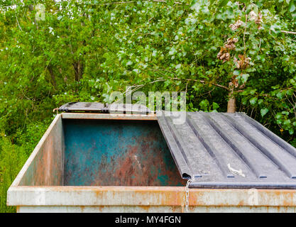 Large empty dirty rusted metal dumpster with half-open lid near trees. Stock Photo