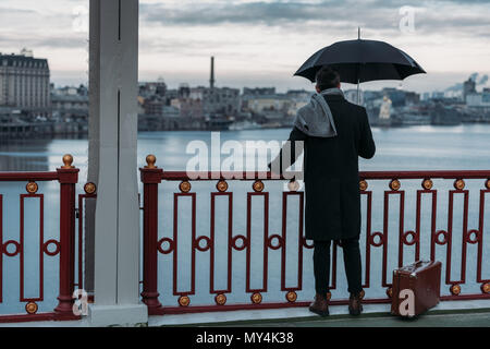 back view of lonely man with umbrella and suitcase standing on bridge Stock Photo