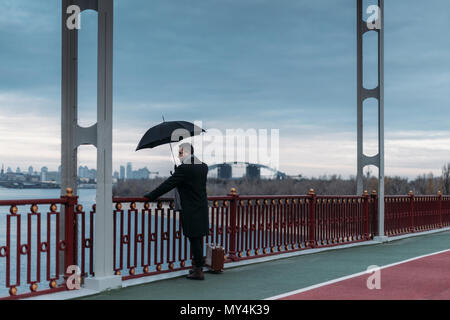stylish lonely man with umbrella and suitcase standing on bridge Stock Photo