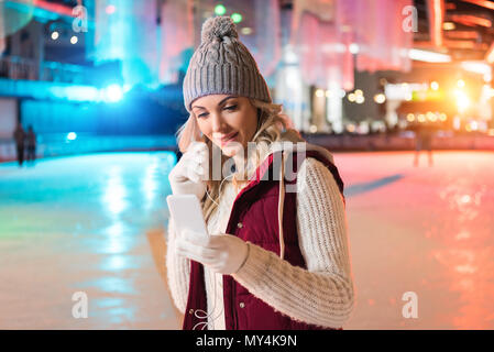 smiling young woman holding smartphone and listening music in earphones on rink Stock Photo