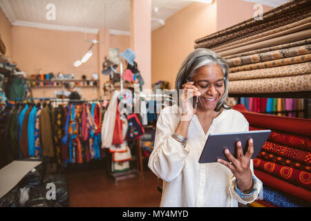 Smiling mature fabric shop owner using a digital tablet and talking on a cellphone while standing by colorful cloths and textiles Stock Photo