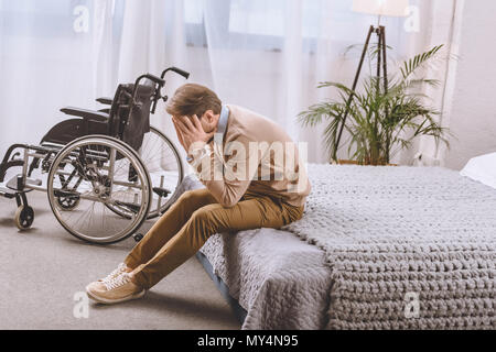 upset man with disability sitting on bed and covering face with hands Stock Photo