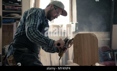 Сarpenter in work clothes and small buiness owner working in woodwork workshop, processes the board with an angle grinder , on the table is a hammer a Stock Photo