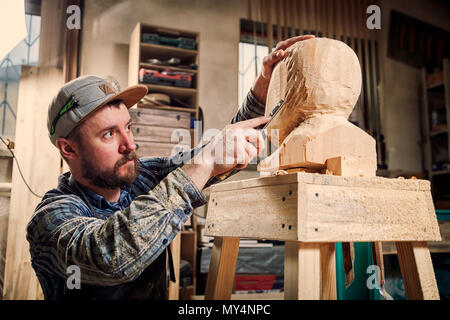 Young man carpenter in a work clothes  saws a man's head with a tree , using a chisel  in the workshop, around a lot of tools for work Stock Photo